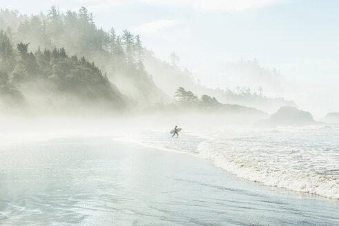 Mann mit Surfbrett, der bei nebligem Wetter am Berg in Richtung Meer läuft - CAVF35683