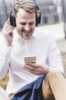 Lächelnder Geschäftsmann mit Kopfhörern und Mobiltelefon - UUF13274