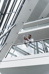 Businessman in office building leaning on railing looking at cell phone - UUF13284