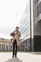 Lächelnder Geschäftsmann mit Mobiltelefon und Basketball vor einem Bürogebäude - UUF13298