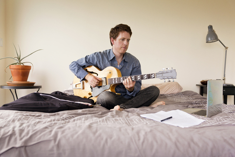 Mann schaut auf Laptop-Computer, während er Gitarre übt, lizenzfreies Stockfoto