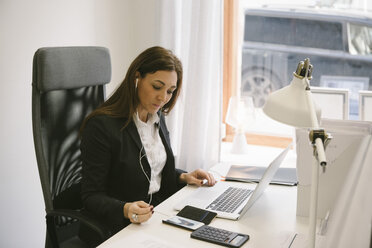 Ältere Immobilienmaklerin hört Kopfhörer, während sie im Büro auf ihren Laptop schaut - MASF02285