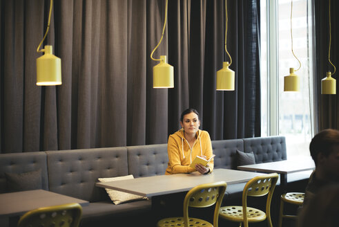 Mid adult businesswoman with book sitting at table in cafeteria at office - MASF02290