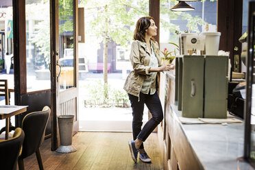 Full length of young woman standing at checkout counter in cafe - CAVF36209