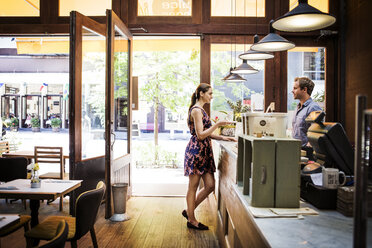 Weiblicher Kunde hält Kaffeetasse, während er mit dem Besitzer am Tresen eines Cafés spricht - CAVF36211
