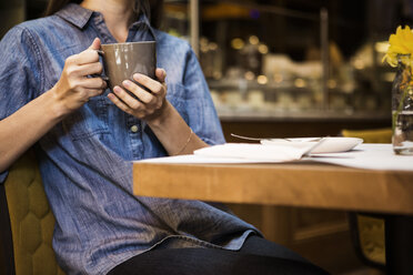 Midsection of woman holding coffee mug sitting at table in cafe - CAVF36216