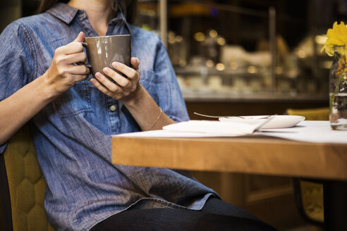 Mittelteil einer Frau, die einen Kaffeebecher hält und am Tisch in einem Café sitzt - CAVF36216