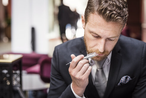 Älterer Geschäftsmann mit Stift im Mund an der Hotelrezeption sitzend - MASF02347