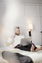 Älterer Geschäftsmann, der einen Laptop benutzt und die Kopfhörer einstellt, während er auf dem Bett im Hotelzimmer sitzt - MASF02349