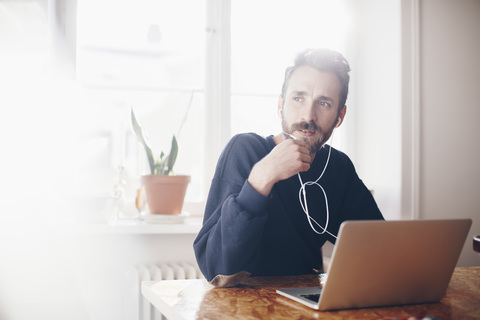 Mann spricht über Kopfhörer am Laptop vor dem Fenster zu Hause, lizenzfreies Stockfoto