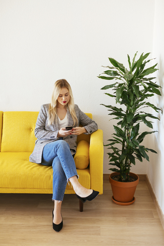 Geschäftsfrau sitzt auf einer gelben Couch und benutzt ein Smartphone, lizenzfreies Stockfoto