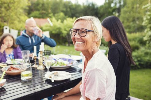 Glückliche Frau sitzt mit Freunden und Familie am Esstisch im Hinterhof während einer Gartenparty - MASF02372