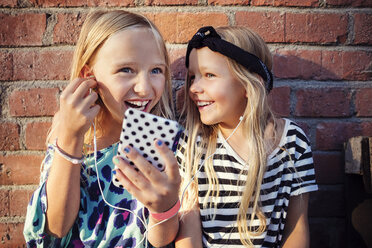 Happy girls listening music through smart phone while sitting against brick wall - MASF02384