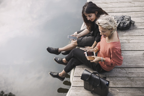 Blick von oben auf glückliche Geschäftsfrauen, die ihr Smartphone benutzen, während sie auf einem Steg am See sitzen, lizenzfreies Stockfoto