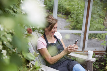 Glückliche Gärtnerin, die ein Mobiltelefon benutzt, während sie im Garten sitzt - MASF02434