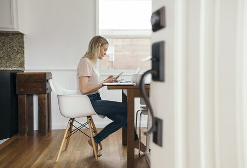 Full length side view of mid adult woman holding mobile phone while using laptop at home - MASF02454
