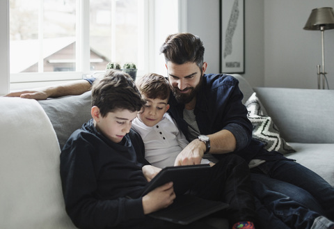 Vater und Söhne benutzen ein Tablet, während sie zu Hause auf dem Sofa sitzen, lizenzfreies Stockfoto