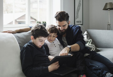 Vater und Söhne benutzen ein Tablet, während sie zu Hause auf dem Sofa sitzen - MASF02467