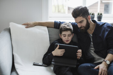 Vater schaut auf einen Jungen, der ein digitales Tablet benutzt, während er auf dem Sofa im Wohnzimmer sitzt - MASF02468
