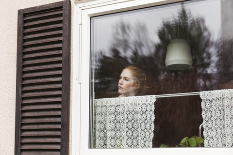 Frau schaut durch ein Fenster, während sie zu Hause steht, lizenzfreies Stockfoto