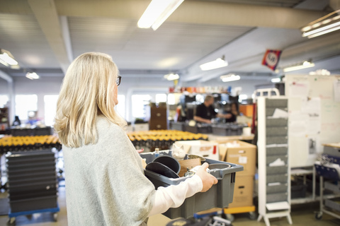 Frau mit blondem Haar trägt Kiste in Werkstatt, lizenzfreies Stockfoto