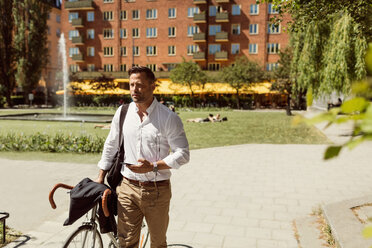 Nachdenklicher Geschäftsmann mit Fahrrad und Smartphone auf dem Fußweg im Sommer - MASF02613