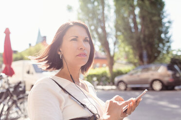 Thoughtful businesswoman looking away while listening music through smart phone on sunny day - MASF02614