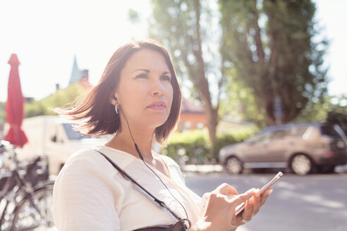 Nachdenkliche Geschäftsfrau, die wegschaut, während sie an einem sonnigen Tag Musik über ihr Smartphone hört - MASF02614