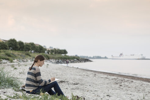 Frau schreibt an einem Buch, während sie am Strand gegen den Himmel sitzt - MASF02616