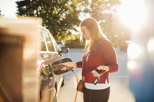 Woman holding smart phone while charging electric car at station on sunny day - MASF02632