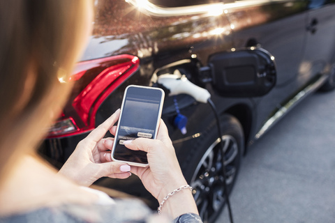 Woman using smart phone while standing by charging electric car stock photo