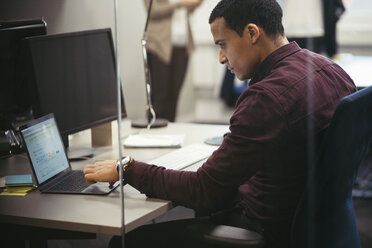 Geschäftsmann mit Laptop am Schreibtisch im Büro mit einem Kollegen im Hintergrund - MASF02721