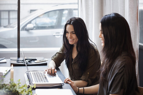 Lächelnde weibliche Kollegen, die einen Laptop benutzen, während sie am Tisch in einem kreativen Büro sitzen - MASF02726