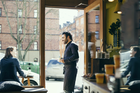 Lächelnder Kellner im Gespräch mit einer Frau auf dem Bürgersteig, gesehen durch Glas in einem Cafe, lizenzfreies Stockfoto