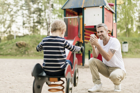 Lächelnder Vater fotografiert seinen Sohn, der auf dem Spielplatz auf einer Federwippe spielt - MASF02743