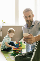 Smiling father using phone while son playing with toy blocks at home - MASF02747