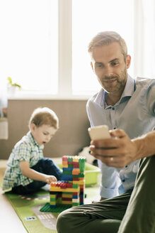 Lächelnder Vater telefoniert, während sein Sohn zu Hause mit Spielzeugblöcken spielt - MASF02747