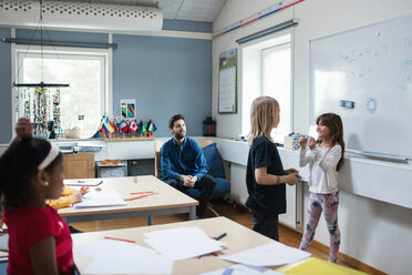 Teacher and students looking at girls discussing while standing against whiteboard - MASF02754