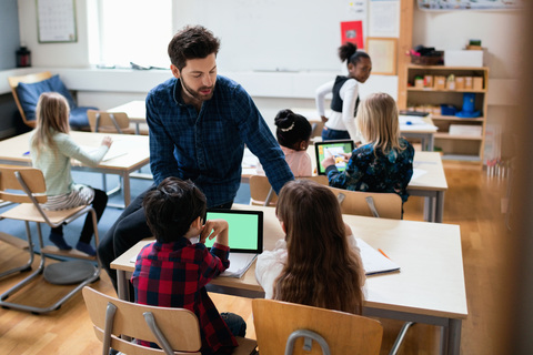 Lehrer sitzt auf dem Schreibtisch und erklärt Schülern die Verwendung eines digitalen Tablets im Klassenzimmer, lizenzfreies Stockfoto