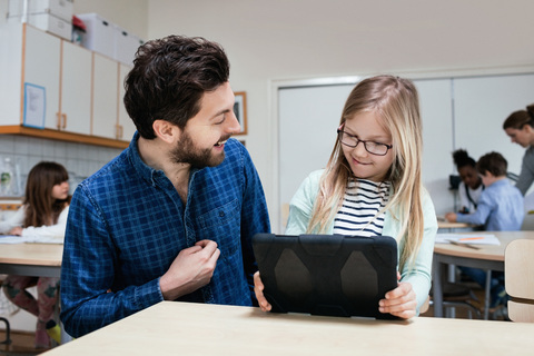 Smiling teacher looking at girl holding digital tablet at desk stock photo
