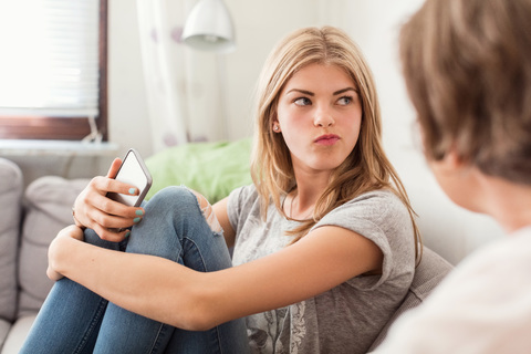 Teenage girl making faces while sitting with mother on sofa at home stock photo