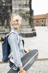 Lächelndes Teenager-Mädchen, das ein Skateboard hält, während es auf einer gepflasterten Straße in der Stadt läuft - MASF02795