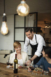 Lächelnder Mann in Schürze serviert Wein an Geschäftsfrauen am Tisch - MASF02806