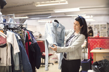 Serious female volunteer looking at jacket by clothes rack in workshop - MASF02856
