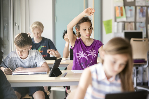 Schulmädchen hebt die Hand am Schreibtisch im Klassenzimmer, lizenzfreies Stockfoto