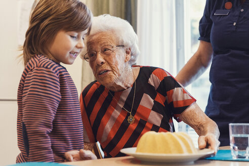 Ältere Frau im Gespräch mit ihrem Urenkel, der einen Biskuitkuchen auf dem Tisch betrachtet, während der Hausmeister zu Hause steht - MASF02890