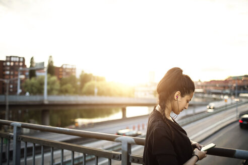 Seitenansicht einer Frau, die ein Mobiltelefon benutzt, während sie auf einer Brücke gegen den Himmel steht - MASF02899