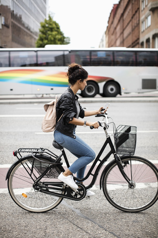 Seitenansicht einer Frau, die ein Mobiltelefon benutzt, während sie mit dem Fahrrad auf einer städtischen Straße fährt, lizenzfreies Stockfoto