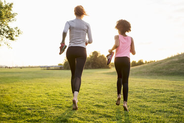 Rear view of woman and girl jogging while holding shoes on grass during sunset - MASF02930