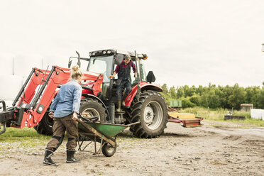 Woman pushing wheelbarrow towards man and tractor at farm - MASF02953